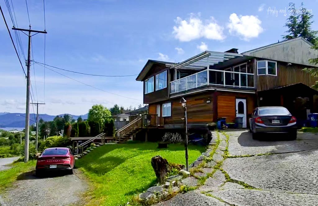 Wooden house with glass-railed balcony and sloped driveway, with two parked cars and mountain views in the background.