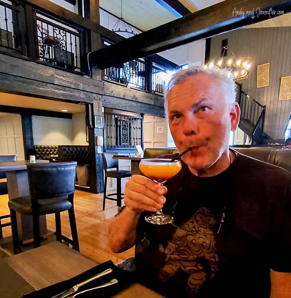Man sipping an orange cocktail through a straw at a cozy, rustic-style restaurant with wooden floors, dark beams, and a loft-style upper level.