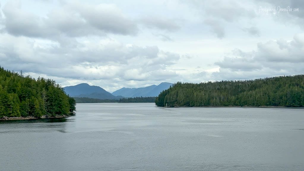 Calm waterway surrounded by forested hills under cloudy sky.