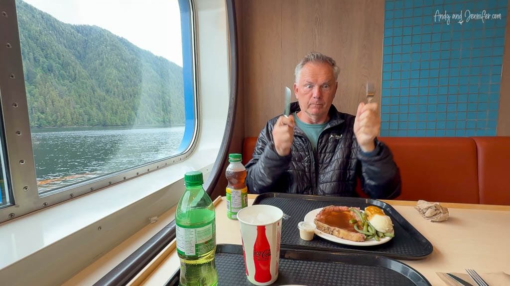 Man holding knife and fork excitedly in ferry dining area with a meal in front of him and a scenic forested view outside the window.
