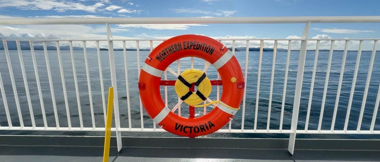 A bright orange life ring labeled "Northern Expedition – Victoria" is mounted on the railing of a ferry deck. The calm ocean and distant landmasses are visible in the background under a partly cloudy blue sky. A green sign next to the ring indicates life-saving equipment.