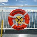 A bright orange life ring labeled "Northern Expedition – Victoria" is mounted on the railing of a ferry deck. The calm ocean and distant landmasses are visible in the background under a partly cloudy blue sky. A green sign next to the ring indicates life-saving equipment.