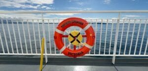 A bright orange life ring labeled "Northern Expedition – Victoria" is mounted on the railing of a ferry deck. The calm ocean and distant landmasses are visible in the background under a partly cloudy blue sky. A green sign next to the ring indicates life-saving equipment.