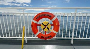 A bright orange life ring labeled "Northern Expedition – Victoria" is mounted on the railing of a ferry deck. The calm ocean and distant landmasses are visible in the background under a partly cloudy blue sky. A green sign next to the ring indicates life-saving equipment.
