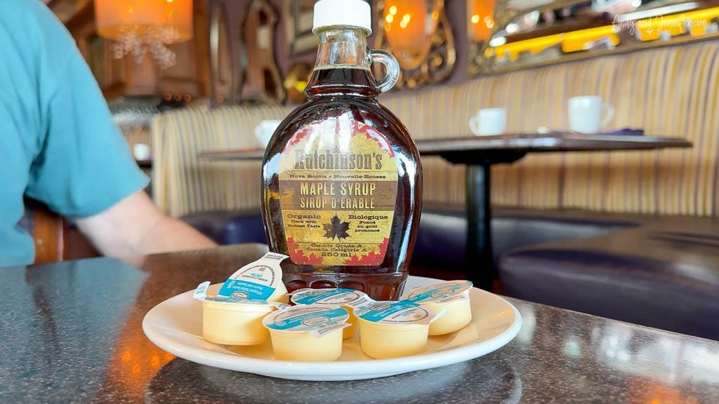 Bottle of Hutchinson’s maple syrup from Nova Scotia on a plate with individual creamers, set on a restaurant table with a cozy booth in the background.