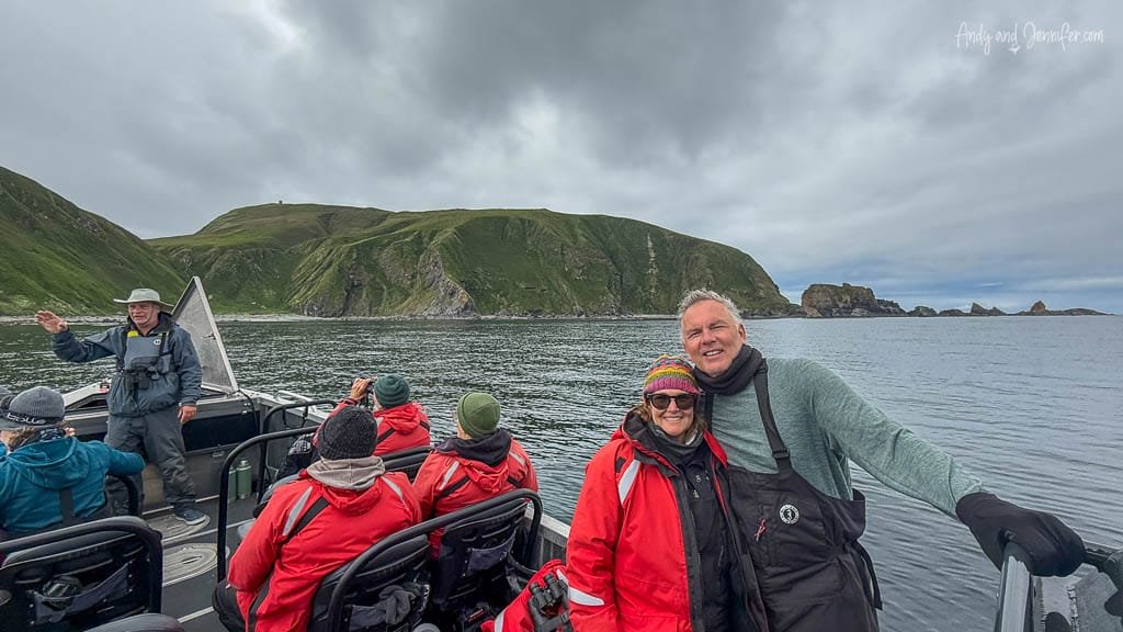Tour group on a boat with a guide, smiling couple in foreground, surrounded by ocean and green cliffs under cloudy skies