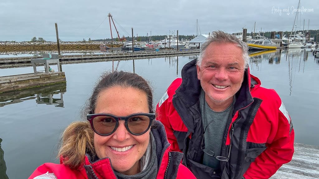 Smiling couple in red jackets taking a selfie at a marina with boats and docks in the background on a cloudy day
