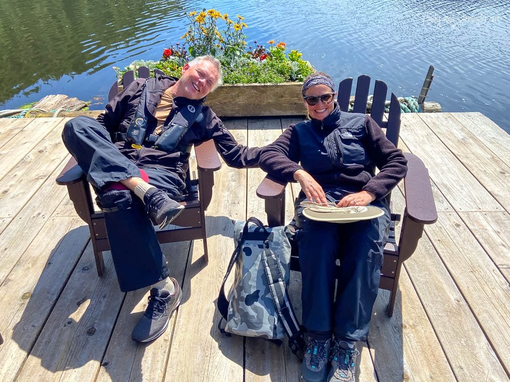 Two people relaxing on wooden deck chairs by the water, holding hands and smiling.