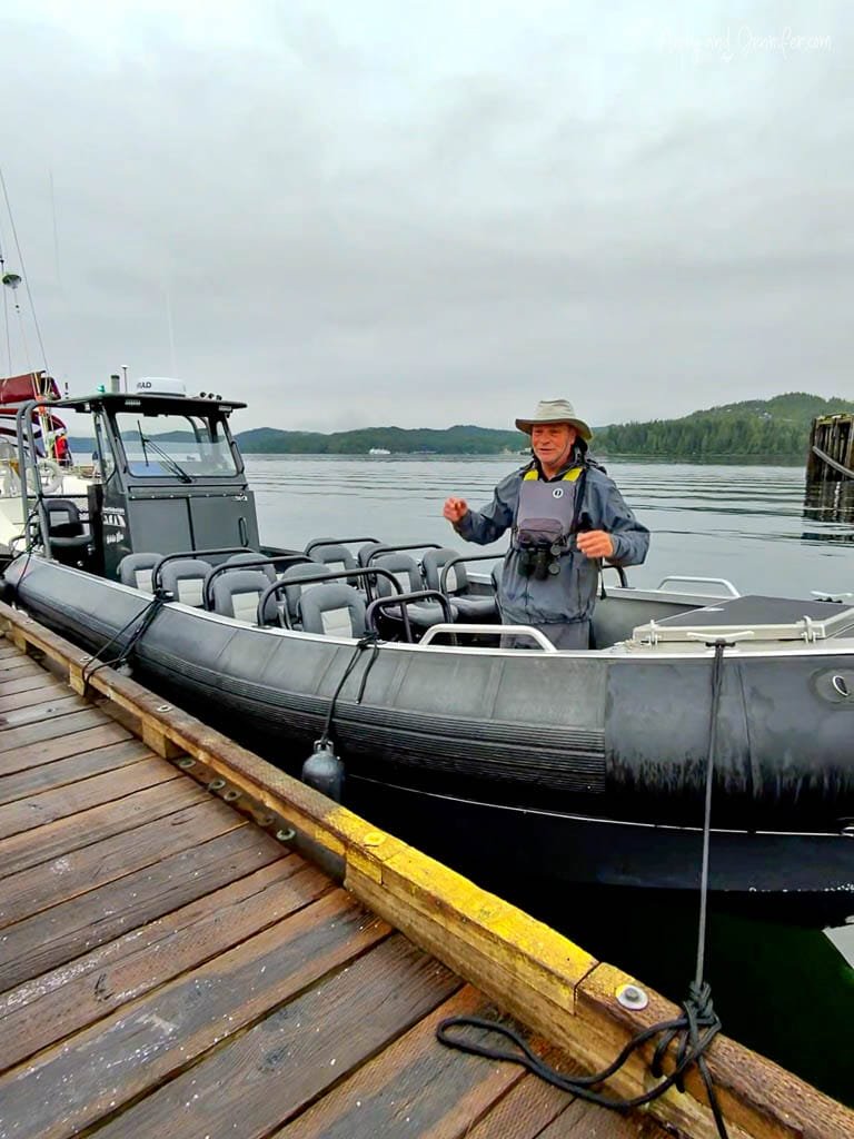 Man in outdoor gear standing on a black inflatable boat docked at a wooden pier on a cloudy day