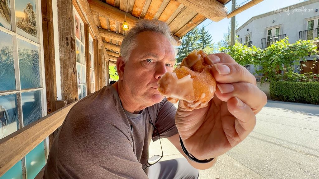 Man holding up a glazed donut hole while sitting outside a rustic wooden building