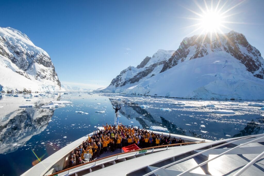 Expedition passengers gathered on the bow of the Quark Ultramarine while cruising through the Lemaire Channel in Antarctica surrounded by mountains, glaciers, and calm icy waters