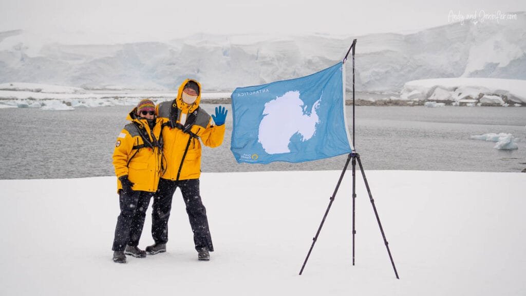 Andy and Jennifer standing beside the Antarctic continent flag during a snowy landing at Portal Point, Antarctica.