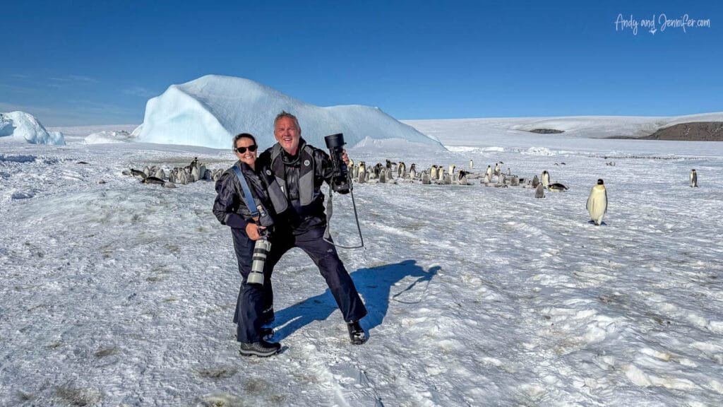 Couple smiling and posing on sea ice near emperor penguins and icebergs in Antarctica