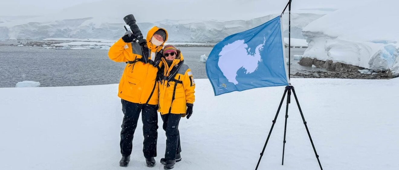 Andy and Jennifer standing beside the Antarctic continent flag during a snowy landing at Portal Point on the Antarctic Peninsula.