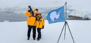 Andy and Jennifer standing beside the Antarctic continent flag during a snowy landing at Portal Point on the Antarctic Peninsula.