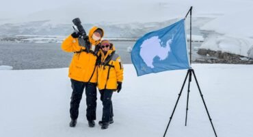 Andy and Jennifer standing beside the Antarctic continent flag during a snowy landing at Portal Point on the Antarctic Peninsula.