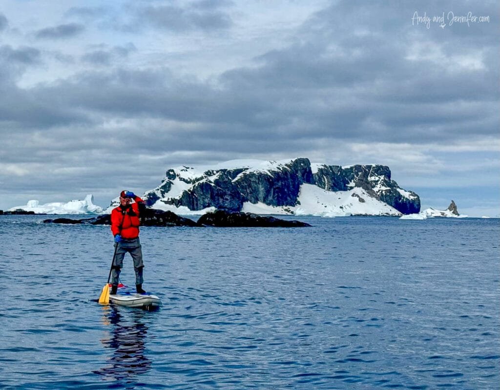 Man stand up paddleboarding in Antarctica near Tower Island surrounded by icebergs, rocky Antarctic islands, and calm polar waters
