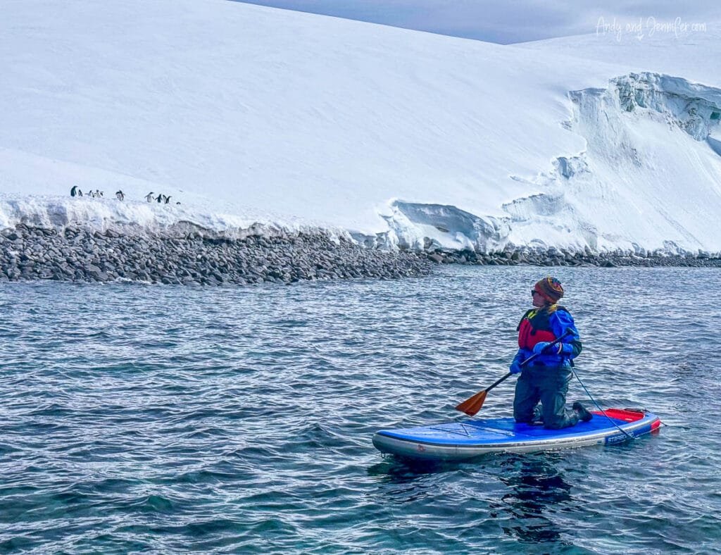 Woman kneeling on a stand up paddleboard in Antarctica near Tower Island with penguins, glaciers, and icy Antarctic coastline in the background