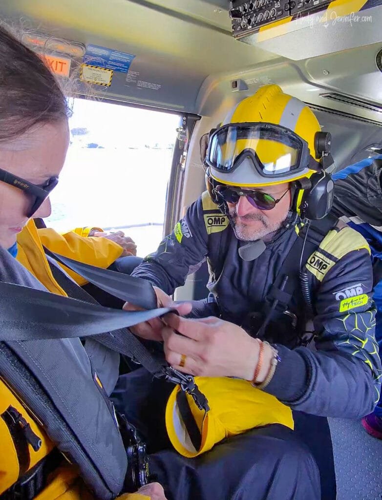 Traveler seated inside helicopter wearing helmet and harness while crew assists before Antarctic flight