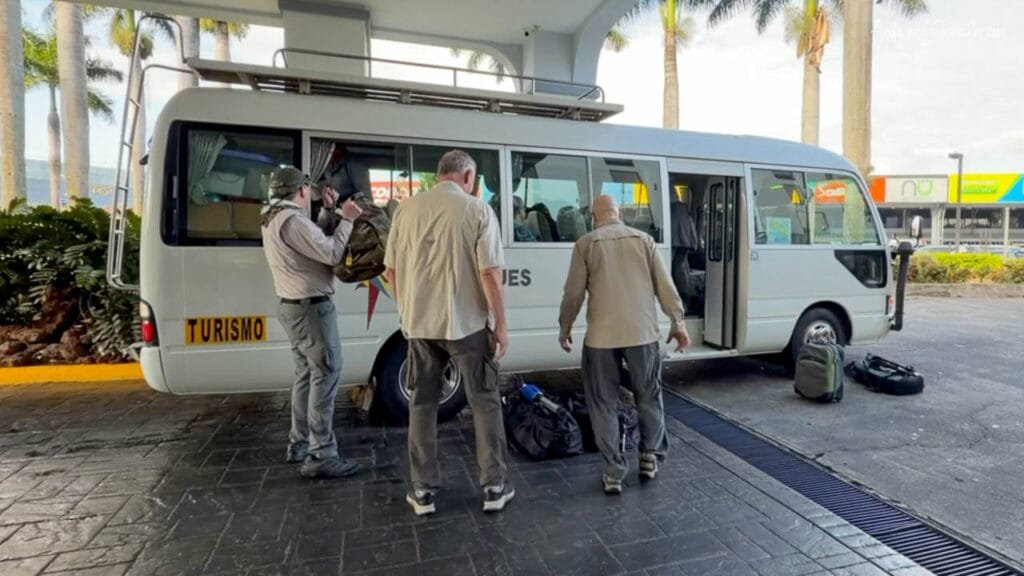 Passengers load camera bags and luggage onto a small white shuttle bus outside a hotel in San José, Costa Rica