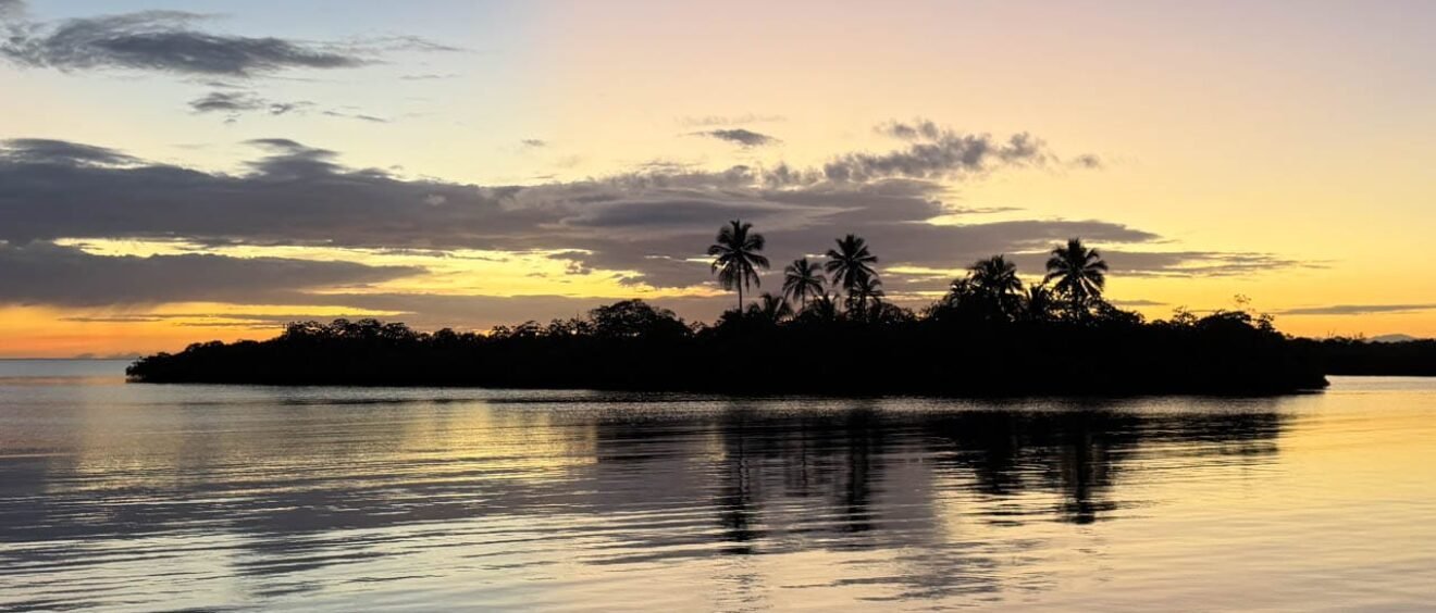 A small tropical island is silhouetted against a golden Caribbean sunset in Panama, with palm trees reflected in the calm water beneath a colourful morning sky.