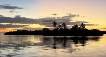 A small tropical island is silhouetted against a golden Caribbean sunset in Panama, with palm trees reflected in the calm water beneath a colourful morning sky.