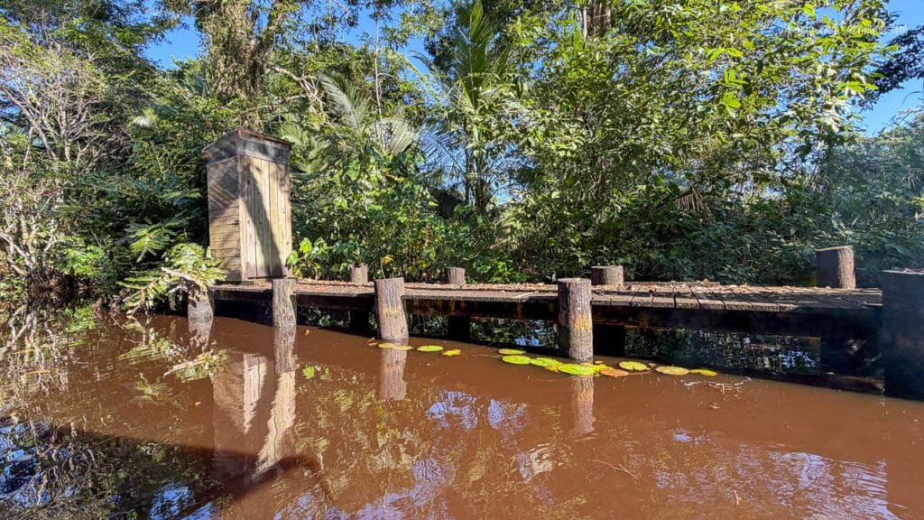 A simple wooden outhouse stands on a small dock beside the Changuinola Canal in Panama, surrounded by dense tropical rainforest and reflected in the calm, tea-coloured water.