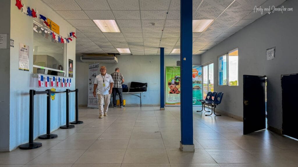 The simple passenger waiting area inside Changuinola Airport in Panama, with a small check-in counter, seating and a few travelers preparing for departure.