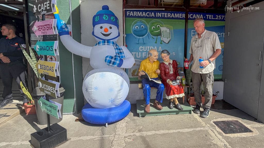 Andy stands outside a roadside convenience store in San José, Costa Rica, beside a whimsical display featuring an inflatable snowman and a statue of an elderly couple reading on a bench while waiting for the shuttle to the airport.