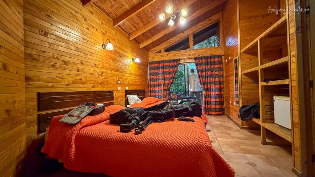 Interior of our wooden cabin at Sueños del Bosque Lodge in Costa Rica's cloud forest, with twin beds covered in camera gear and luggage, and Andy standing on the balcony overlooking the forest.