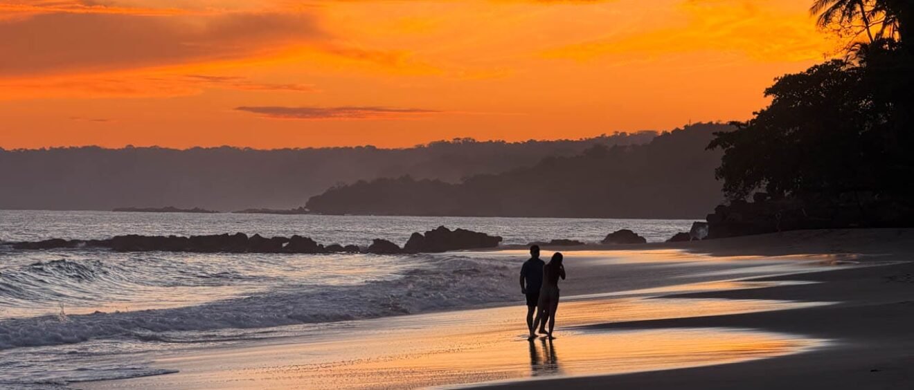 Couple walking along the beach at sunset at Tango Mar Beach Resort, Costa Rica, beneath a vivid orange sky.
