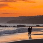 Couple walking along the beach at sunset at Tango Mar Beach Resort, Costa Rica, beneath a vivid orange sky.