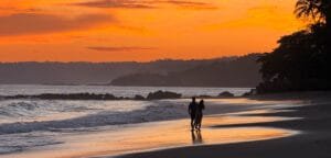 Couple walking along the beach at sunset at Tango Mar Beach Resort, Costa Rica, beneath a vivid orange sky.