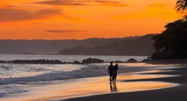 Couple walking along the beach at sunset at Tango Mar Beach Resort, Costa Rica, beneath a vivid orange sky.