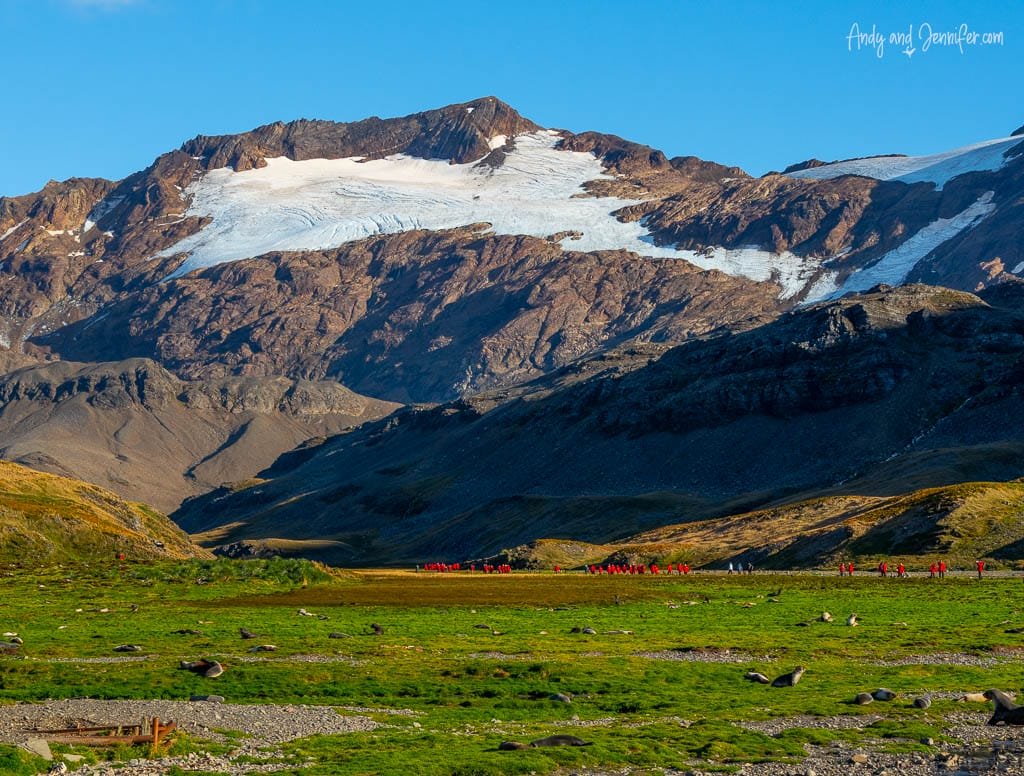 Hikers crossing the grassy valley beneath snow-covered mountains and glaciers at Stromness, South Georgia.