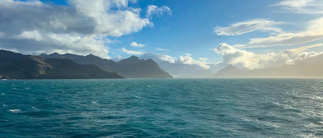Mountainous coastline and turquoise waters of Jason Harbour on South Georgia under clearing skies.